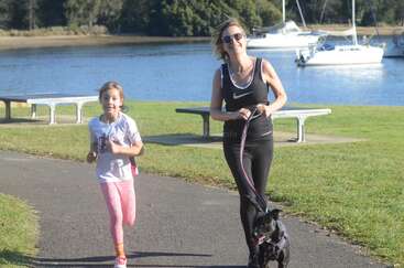 A woman and a young girl are jogging on a sunny day by a river, with a black dog on a leash. Boats are docked nearby.