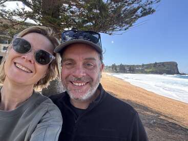 A smiling couple wearing sunglasses poses for a selfie on a sunny day at a beautiful sandy beach, with waves, trees, and cliffs in the background.