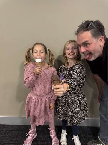 Two young girls enjoy ice cream cones, one in a pink dress and boots, the other in leopard print. A playful man leans in, sticking out his tongue.