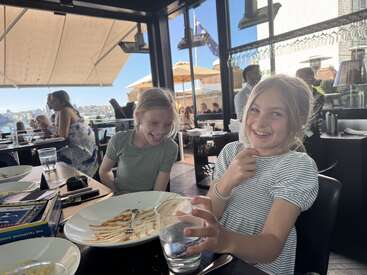 Two young girls laugh and smile at a restaurant table with empty plates. Sunlight streams in through windows, with other diners and outdoor seating visible.