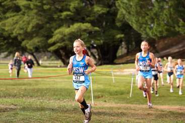 Young athletes participate in a cross-country race, running through a grassy park area with trees in the background. The lead runner is wearing a blue NSW uniform.