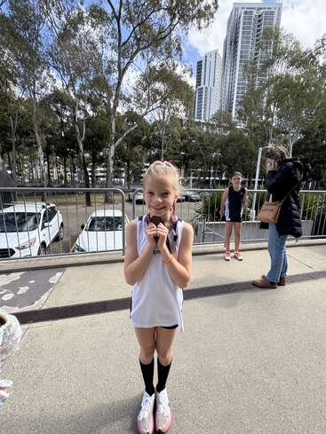 A young girl in sports attire proudly displays a medal outdoors. Tall buildings, trees, two other people, and parked cars are visible in the background. Sunny day.