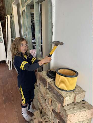 A young child with long hair holds a hammer, poised to strike a white wall. Bricks and a yellow filter sit nearby; construction environment.