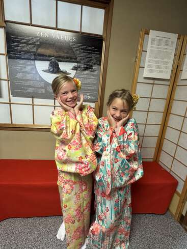 Two smiling girls dressed in colorful floral kimonos pose cheerfully indoors, standing in front of a poster titled "Zen Values" and traditional Japanese decor.