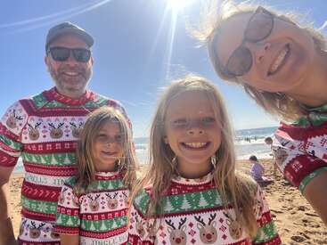 A smiling family of four, all wearing festive Christmas shirts, poses together on a sunny beach. The ocean and blue sky are visible behind them.