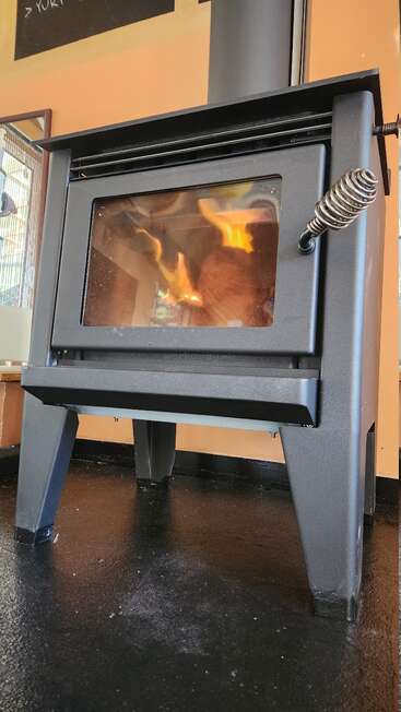 A modern black wood-burning stove with a glass door, showing a warm fire inside. The stove stands on sturdy legs on a dark floor.