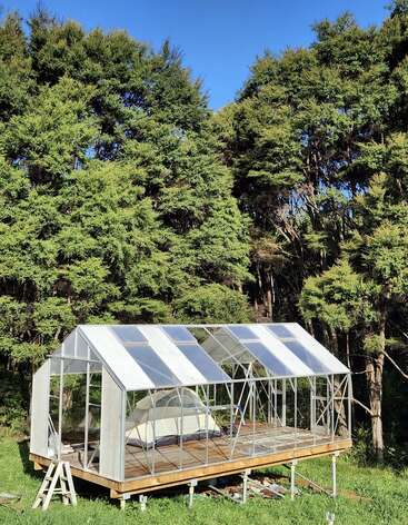 A partially constructed greenhouse stands on a raised platform amidst lush green grass, surrounded by dense, tall trees, beneath a clear, bright blue sky.