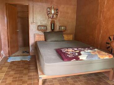 This cozy room features brown walls, a double bed with pillows, a floral blanket, a small woven rug, tiled floor, and an open door to another space.