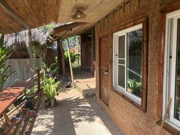 A rustic porch with a bamboo roof, clay walls, wooden-framed windows, plants, a long bench, and abundant sunlight, creating a warm, inviting, tropical atmosphere.
