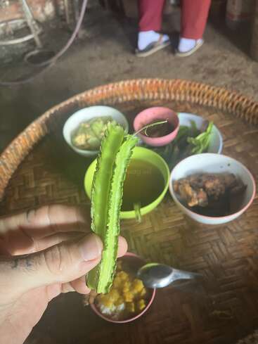 A hand holds a green winged bean above a woven tray with several colorful bowls containing various foods. Someone wearing sandals and red pants sits in the background.