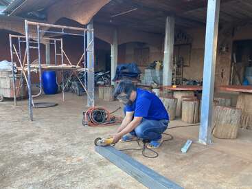 A person wearing safety goggles and a blue shirt is cutting a metal beam with a power tool outdoors. Scaffolding, bikes, and tools are in the background.