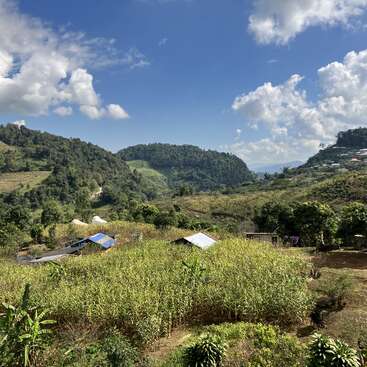 Green hills surround a rural landscape with cornfields, scattered small houses, and lush trees under a bright blue sky filled with fluffy white clouds. Peaceful scenery.