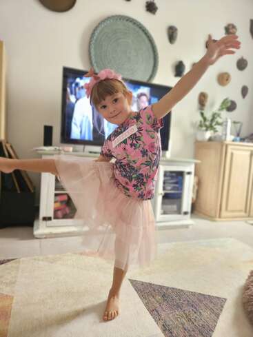 The image depicts a young girl in a pink floral shirt and tutu, standing on one leg with her arms outstretched, in a living room with a TV and various decorations on the wall.