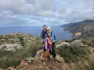 A family of three and their dog pose on a rocky hilltop with scenic ocean views, surrounded by green shrubs, under a cloudy sky.