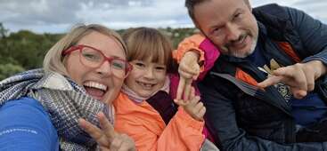 A joyful family of three poses closely together outdoors, smiling brightly. The woman wears red glasses, the child an orange jacket, and everyone gestures peace signs.