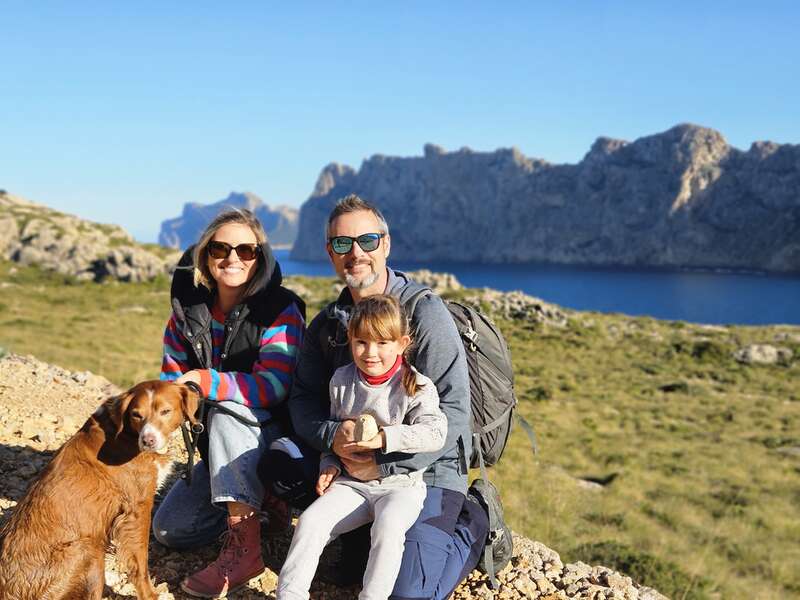 The image depicts a family of three, accompanied by a dog, posing for a photo on a rocky outcrop overlooking a serene body of water and majestic mountains.