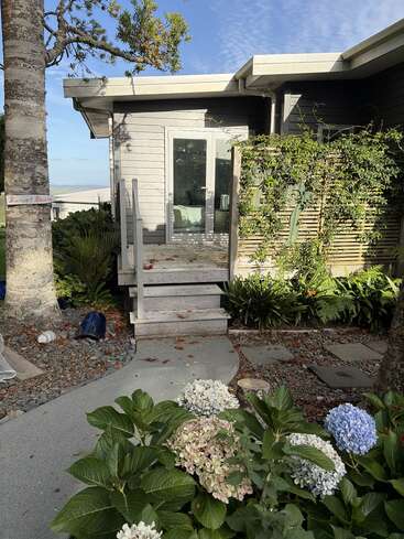 A small house with glass doors and stairs, surrounded by green plants, colorful hydrangeas, gravel path, and a sign reading "Sunset Studio" on a tree.