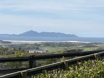 The image shows a scenic coastal landscape with lush green fields, a sandy beach, ocean waters, distant island mountains, and a wooden fence in the foreground.