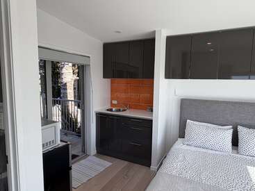 This image shows a compact studio apartment with a modern kitchenette, a small bed with polka-dot bedding, a balcony with sliding doors, and natural light.