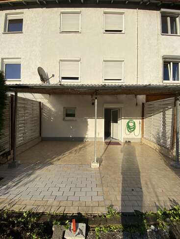 Ein dreistöckiges Gebäude mit weißen Wänden hat eine geflieste Terrasse, eine Metallmarkise, einen Gartenschlauch, eine Satellitenschüssel und geschlossene Fensterläden; ein langer Schatten ist zu sehen.
