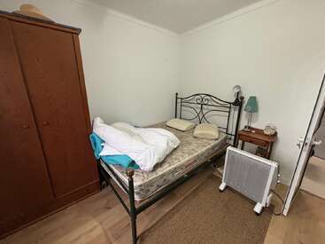 A small, simple bedroom with a metal bed frame, bare mattress, pillows, wardrobe, side table, heater, lamp, and a white wall background. Minimal decor.