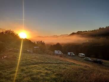 L'image montre un magnifique lever de soleil sur une vallée brumeuse, avec la lumière du soleil qui traverse les champs herbeux, les collines boisées, les voitures et les camping-cars garés en contrebas.