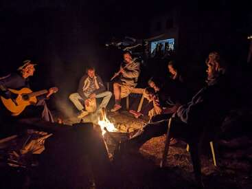 Un groupe de personnes assises autour d'un feu de camp la nuit, jouant de la guitare, de la flûte et des percussions, appréciant la musique et la conversation à la lueur chaleureuse du feu.
