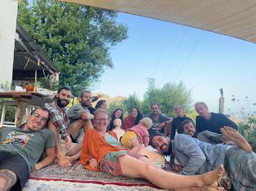 Un groupe de personnes se détend en plein air sur un tapis à motifs, à l'ombre. Elles sont souriantes, les arbres et les montagnes étant visibles à l'arrière-plan.
