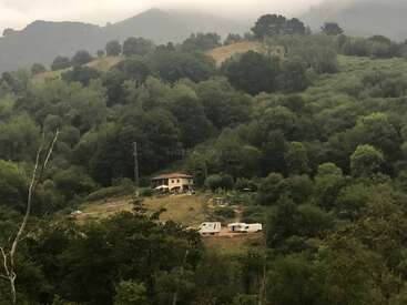 Une petite maison se trouve sur le flanc d'une colline entourée d'une forêt dense et verte, avec deux caravanes blanches garées à proximité. Des montagnes brumeuses s'élèvent à l'arrière-plan. Paysage paisible.