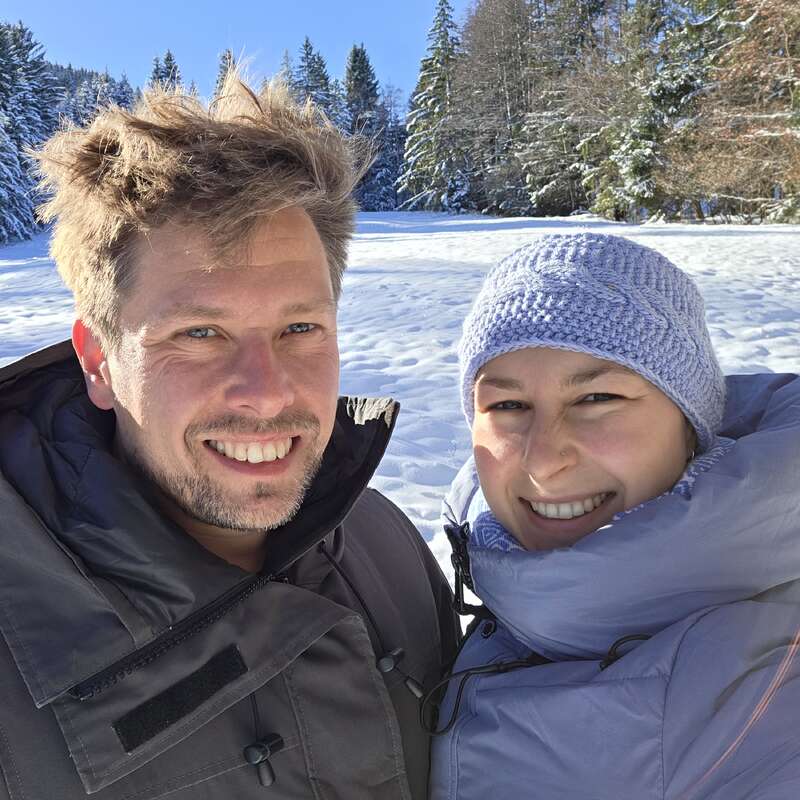 A smiling couple dressed warmly poses together outdoors on a sunny, snowy day. Pine trees and a clear blue sky create a picturesque winter background behind them.