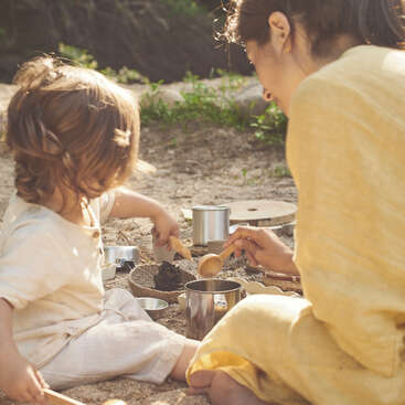 A woman and a young child play together outdoors in the sand, using wooden spoons and metal containers, enjoying a warm, sunlit afternoon in nature.