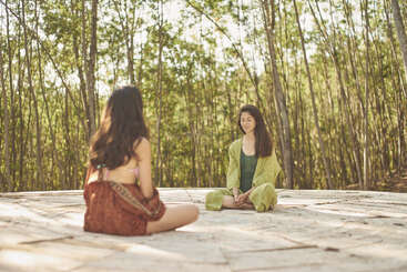 Two women are sitting cross-legged on a stone platform in a peaceful forest setting, facing each other, appearing relaxed and engaged in meditation or conversation.