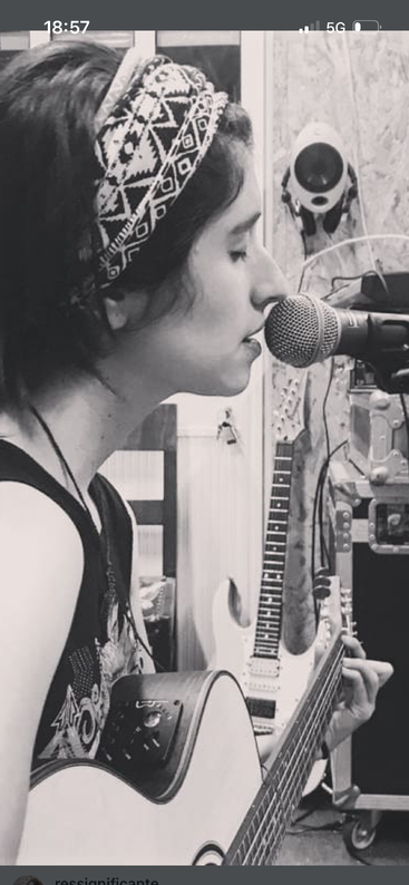 A person with a patterned headband sings into a microphone, playing an acoustic guitar. Music equipment and an electric guitar are visible in the background. Black and white.