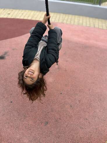 A child in a black jacket smiles joyfully while hanging upside down from a rope at a playground, with a red and yellow surface beneath them.
