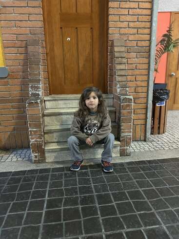 A young child with long hair sits on stone steps in front of a wooden door, surrounded by brick walls, looking calmly at the camera.