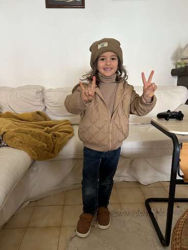 A smiling child stands indoors wearing a brown beanie, tan quilted jacket, jeans, and brown shoes, flashing peace signs with both hands, on tiled flooring.