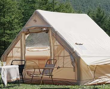 A large beige tent stands amidst lush green trees, with two chairs and a table set outside, creating a cozy and inviting outdoor camping scene.