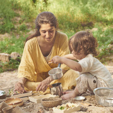 A woman and a young child play together outdoors, sitting on sandy ground, surrounded by baskets, kitchen utensils, and leaves, enjoying a nature-inspired activity.
