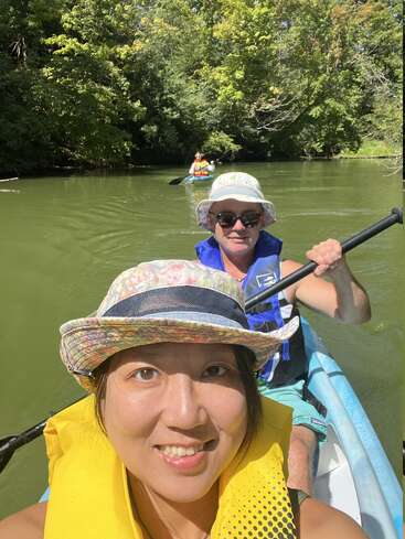 A smiling woman takes a selfie while kayaking with another person on a green river surrounded by lush trees, both wearing hats and life jackets, enjoying nature.