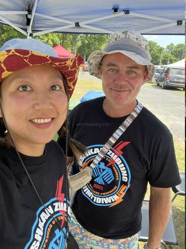Two people wearing hats and matching black event t-shirts stand under a tent outdoors, smiling at the camera. Trees and parked cars are visible behind them.