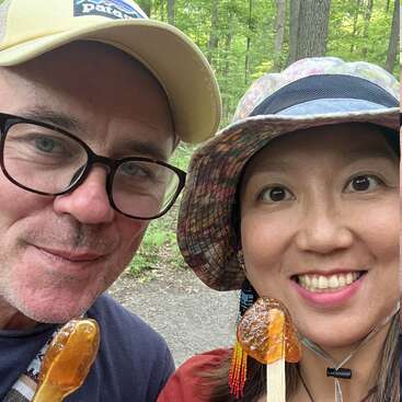 A smiling man and woman take a close-up selfie outdoors, each holding maple taffy sticks, wearing hats and surrounded by green forest scenery. They look happy.