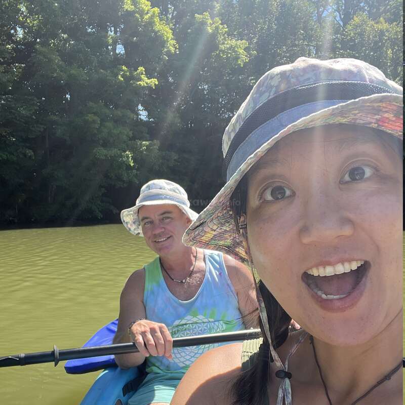 Two people in sun hats are kayaking on a river. The woman in front is smiling excitedly, while the man behind her paddles. Lush green trees surround them.