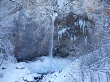 A tall, narrow waterfall cascades down a rocky cliff, surrounded by snow-covered trees and large icicles, creating a breathtaking, icy winter landscape.