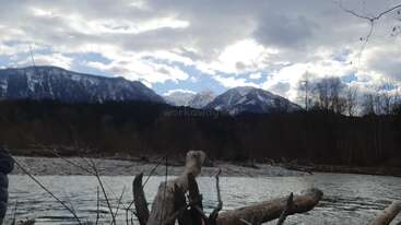 A peaceful river flows in the foreground with driftwood scattered along the shore. Snow-capped mountains and cloudy sky dominate the serene, natural landscape.