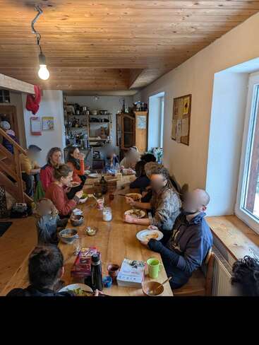A group of people, adults and children, sit around a wooden table sharing a meal in a cozy kitchen with natural light, enjoying food and conversation.