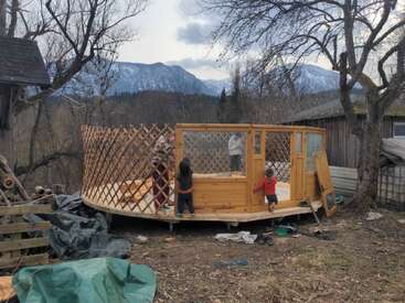 Eine teilweise fertiggebaute Jurte steht draußen auf Holzstützen. Drei Kinder und ein Erwachsener bauen den Holzrahmen auf, umgeben von Bäumen und schneebedeckten Bergen in der Ferne.