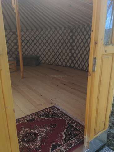 La imagen muestra la entrada de madera de una yurta con una alfombra de motivos rojos, suelo de madera, paredes de celosía y un mobiliario interior minimalista, todo ello bañado por la luz natural.