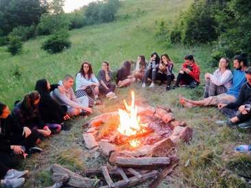Um grupo de pessoas está sentado em círculo ao redor de uma fogueira num campo gramado, curtindo a natureza, conversando e relaxando juntos durante uma noite ao ar livre.