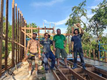 Quatro homens, felizes e sorridentes, estão em um canteiro de obras de madeira, cercados por árvores e sob um céu azul, parecendo estar trabalhando juntos em um projeto de construção.