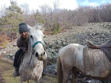 Uma pessoa sorridente, usando um chapéu cinza, monta um cavalo branco com um cabresto azul. Outro cavalo selado está parado ali perto. Árvores sem folhas e rochas preenchem o fundo.